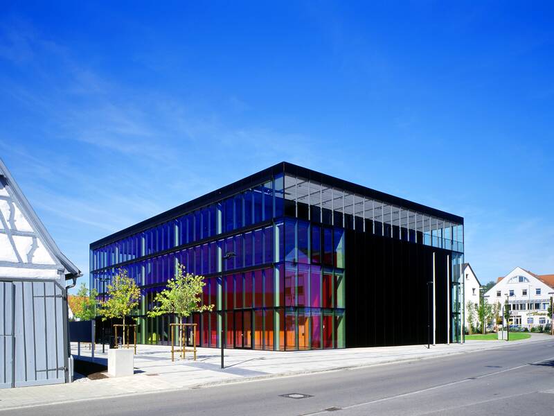 Das Uditorium mit seiner markanten Glasfassade im Sonnenschein und vor blauem Himmel. Es steht an einer grauen und leeren Straße. Davor befinden sich Bäume und daneben ist ein Teil eines Fachwerkhauses mit grauen Balken zu sehen.