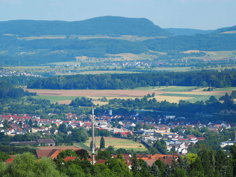 Eine Sicht auf Uhingen von einem Aussichtspunkt aus. Im Vordergrund Gebäude in Miniaturansicht und dichte, grüne Bäume, hinten die gelbe Getreidefelder und die Berge der Voralb