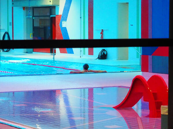Eine Person lehnt sich im Becken des Hallenbads an, davor ist die rote Rutsche im Planschbecken.