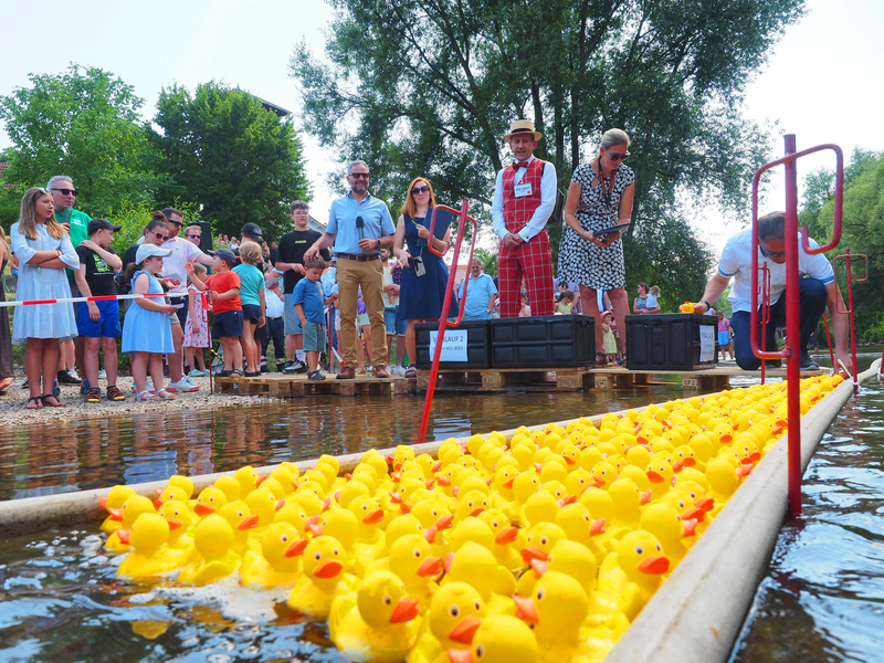 Auf der Fils schwimmen zahlreiche gelbe Gummienten, links am Ufer schauen viele Menschen dem Entenrennen beim Kandelhock zu.