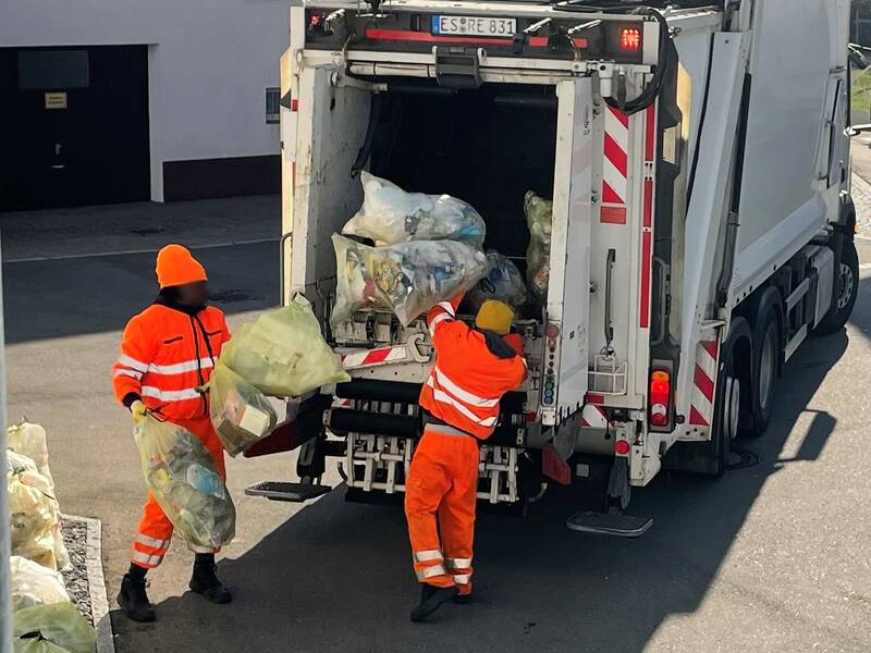 Zwei Männer in orangefarbener Kleidung werfen gelbe Plastiksäcke in die Rückseite eines Müllfahrzeugs, an dessen Heck sie stehen.