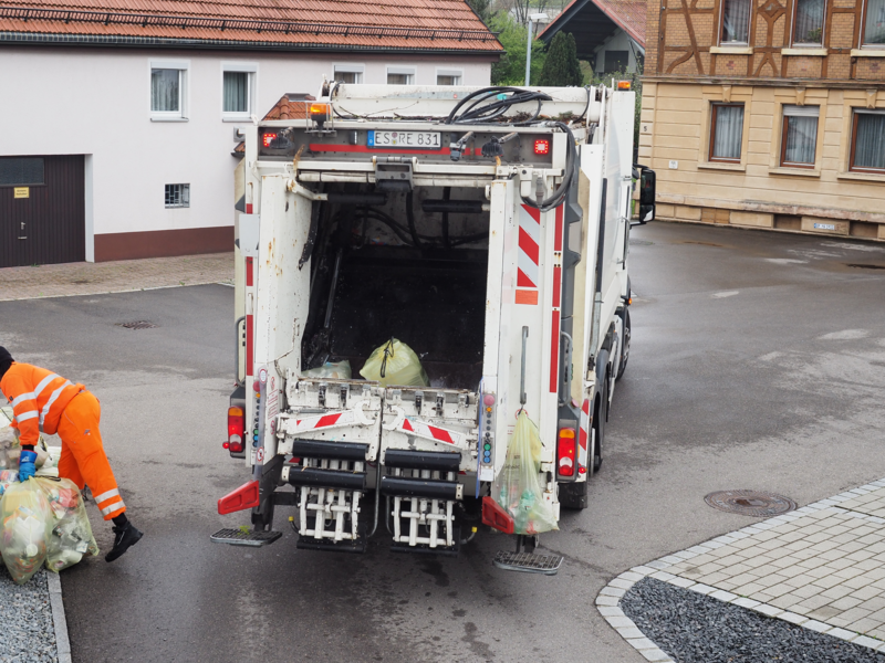 Ein Müllmann in orangefarbener Kleidung beugt sich zu einigen gelben Plastiksäcken, rechts von ihm ist die Rückseite eines Müllfahrzeugs. Dort kommen die gelben Säcke dann rein.