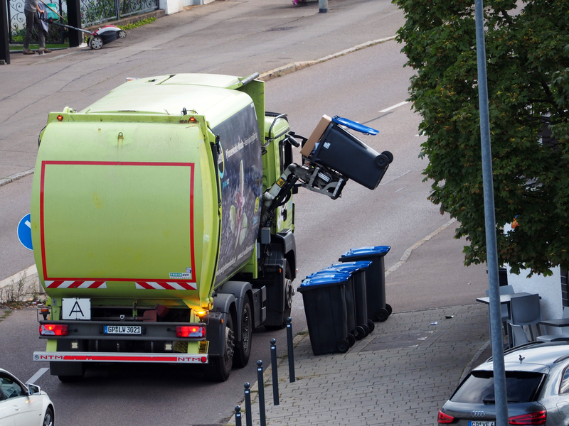 Ein grünes Müllfahrzeug hebt eine schwarze Mülltonne mit blauem Deckel und führt sie zu einer Öffnung an der Seite des Wagens, um die Tonne zu entleeren.