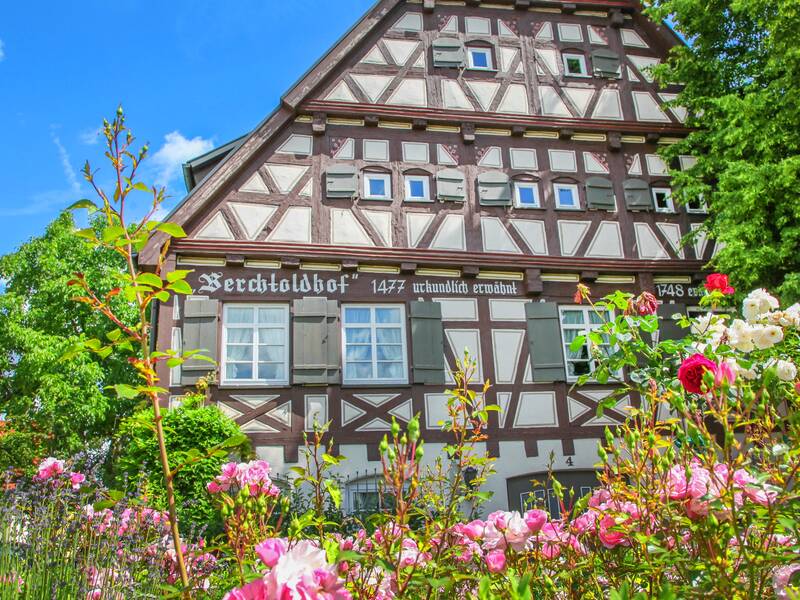 Blühende Blumen vor dem historischen Fachwerkhaus Berchtoldshof, an dessen Fassade braune Balken und grüne Fensterläden zu sehen sind.