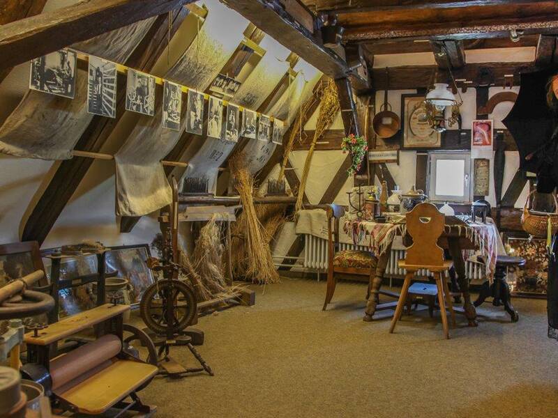 Das Innere des Stadtmuseums: Historische Arbeitsgeräte aus dem Alltag der Menschen wie ein Spinnrad stehen am Boden, an der schrägen Decke hängen Bilder und rechts steht eine Schaufensterpuppe, die ein schwarzes Kleid samt schwarzer Haube trägt.