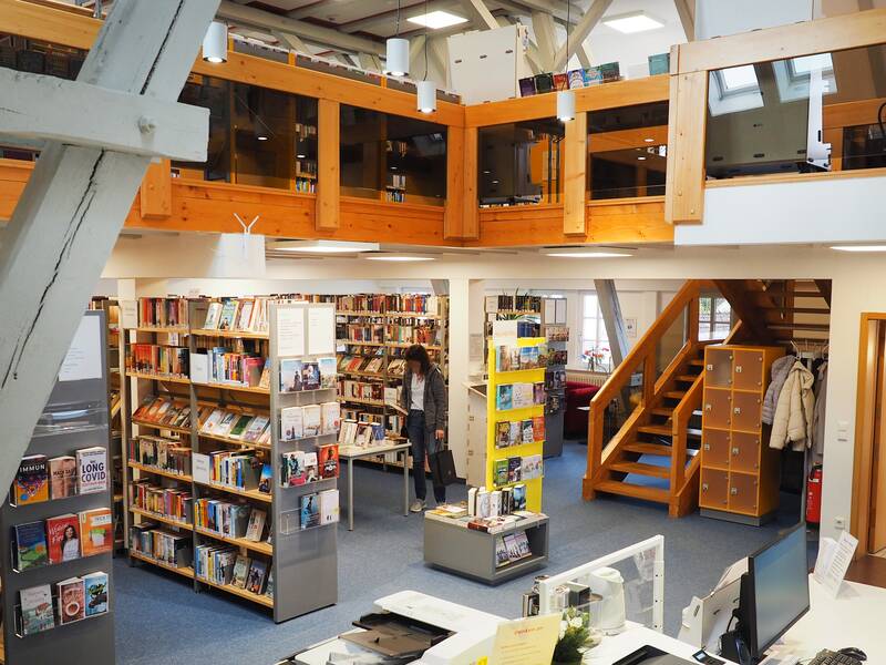 In der Stadtbücherei stehen mehrere Regale voller Bücher auf einem grauen Teppich. Darüber ist eine weitere Etage mit einem Holz-Glas-Geländer. 