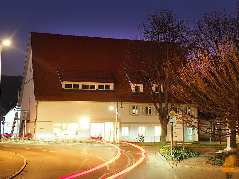 Das K1 in der blauen Stunde am Abend, wenn der Tag endet und die Nacht beginnt. Im Erdgeschoss leuchten Lampen und die Straßenlaternen leuchten ebenfalls hell. Von vorbeifahrenden Autos sind die strahlenden Lampen in rot und gelb zu sehen.