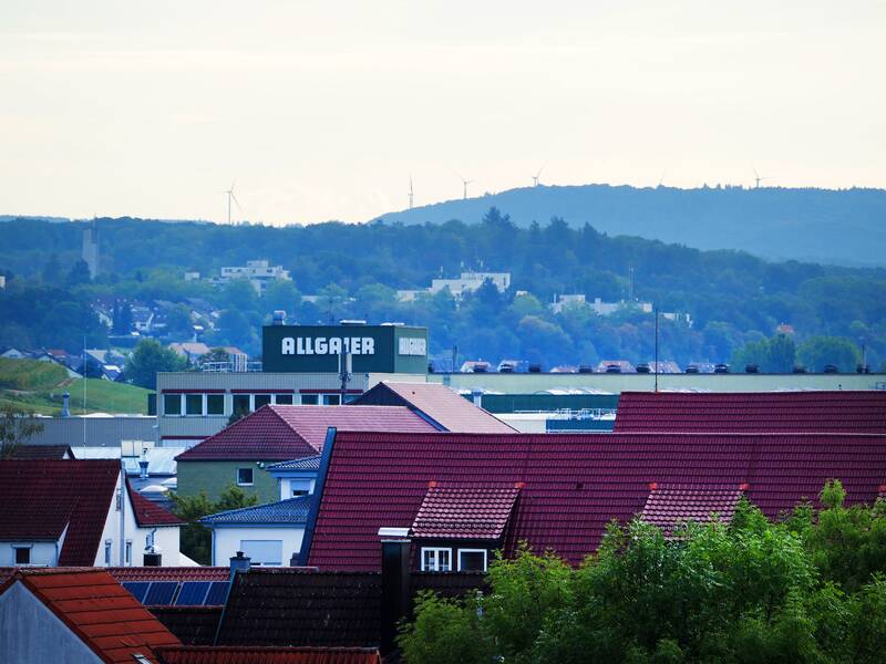 Blick über Uhingen mit roten Dächern im Vordergrund, einer Fabrik mit dem Schriftzug Allgaier und  Wald sowie Windräder am Horizont.
