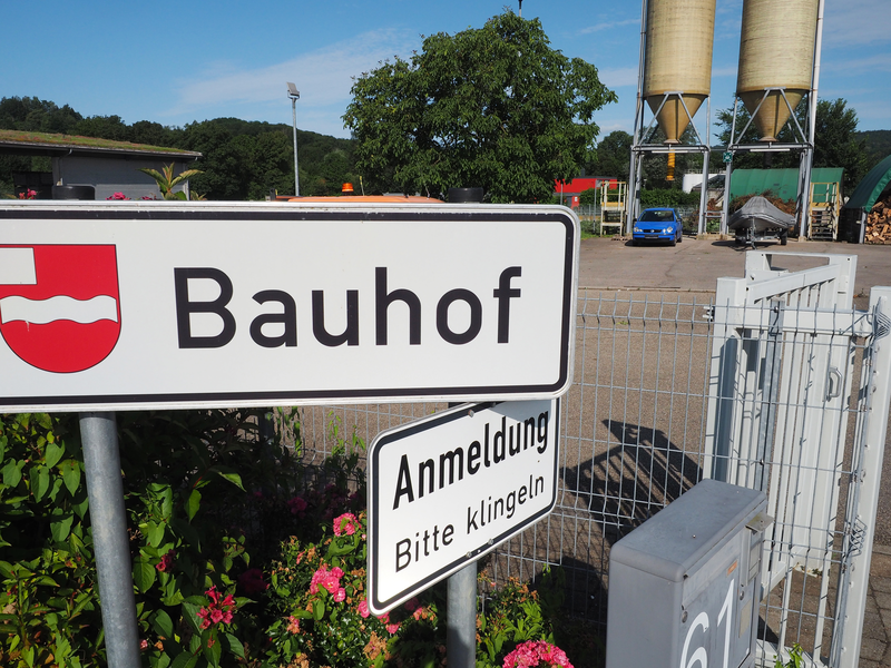 Ein Schild mit der Aufschrift "Bauhof" und einem kleineren Schild darunter mit "Anmeldung, Bitte klingeln" steht vor einem Zaun mit einem Parkplatz und Silos im Hintergrund.