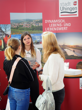 Am Stand der Stadt Uhingen - rote Hintergrundfarbe und Ansichten von Uhingen - stehen zwei junge Frauen, die von hinten zu sehen sind. Sie sprechen mit einer braunhaarigen Stadtangestellten.