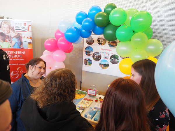 An einem Stand sind mehrere Frauen in ein Gespräch vertieft. Im Hintergrund ist ein Regenbogen aus bunten Luftballons geformt. 