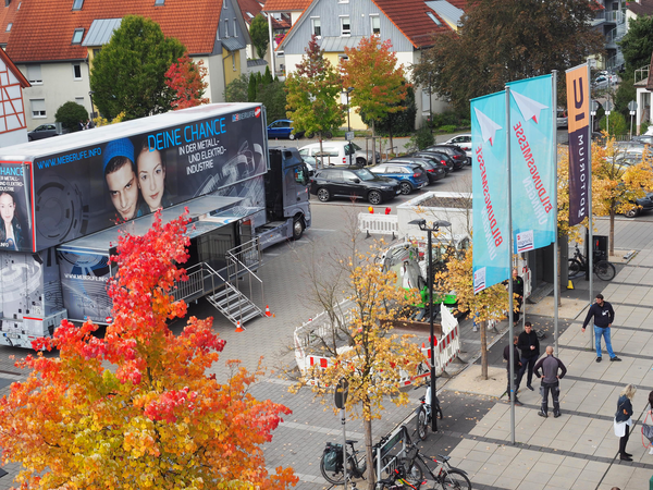 Auf dem Parkplatz vor dem Uditorium steht ein großer Lastwagen. Dessen Anhänger ist doppelt so hoch wie gewöhnliche Anhänger. In dem Messe-Truck wird über Berufe aus der Metall- und Elektroindustrie informiert. Im Vordergrund  sind bunt verfärbte Bäume und Fahnen zu sehen. Die Fahnen sind türkis und schwarz-orange.