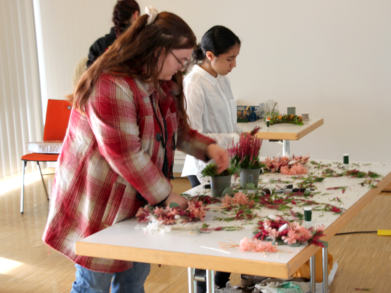 Junge Frauen binden in einem Workshop bei der Bildungsmesse Blumenkränze mit Material, das vor ihnen auf einem Tisch liegt.