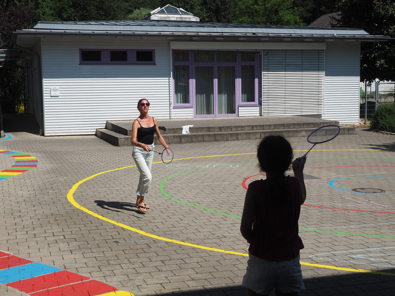 Auf einem Schulhof spielen ein Mädchen und eine Betreuerin Federball.