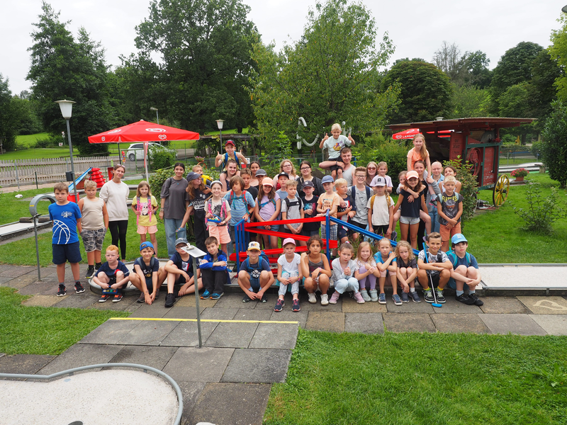 Fast 40 Mädchen und Jungen stehen und sitzen in mehreren Reihen auf einer Mini-Golf-Anlage und winken fröhlich.