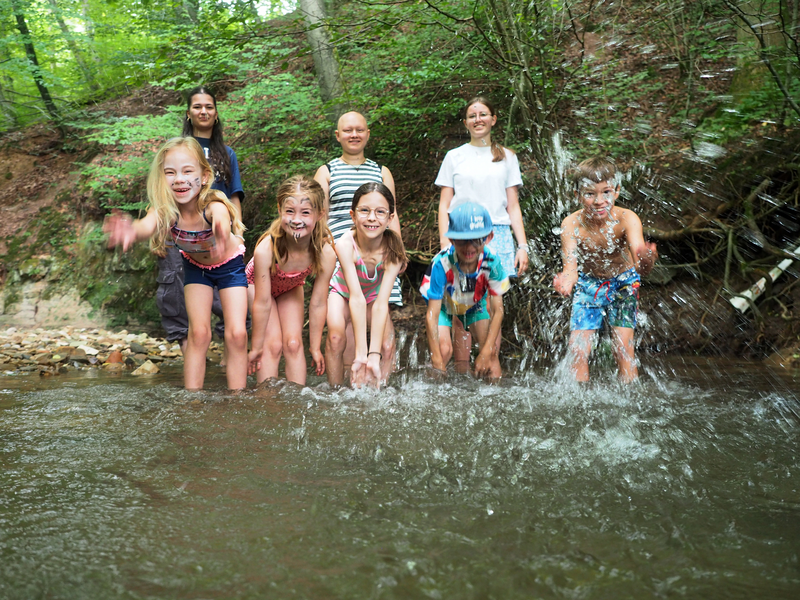 Fünf Kinder stehen knöcheltief im Wasser eines Baches und spritzen mit Wasser um sich. Hinter ihnen stehen drei Betreuerinnen.