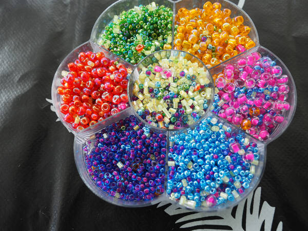 In einem durchsichtigen Behälter in Form einer Blüte liegen zahlreiche bunte Perlen. In jedem Blütenblatt eine andere Farbe.