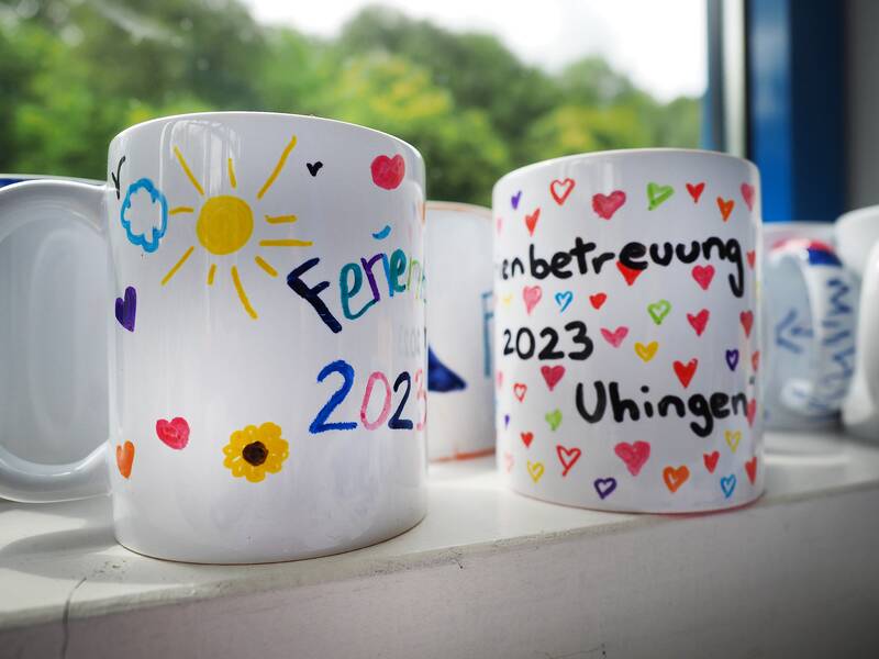 Von Kindern bemalte Tassen stehen auf einem Fensterbrett. Auf den weißen Tassen sind bunte Herzen, eine Sonne und der Schriftzug "Ferienprogramm 2023".