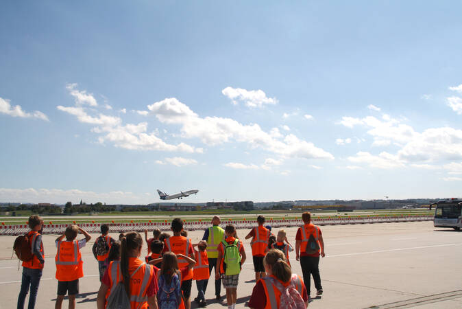 Kinder in orangefarbenen Warnwesten stehen auf dem Flughafen Stuttgart auf einem Rollfeld und sehen in der Ferne, wie ein Flugzeug in den blauen Himmel abhebt.