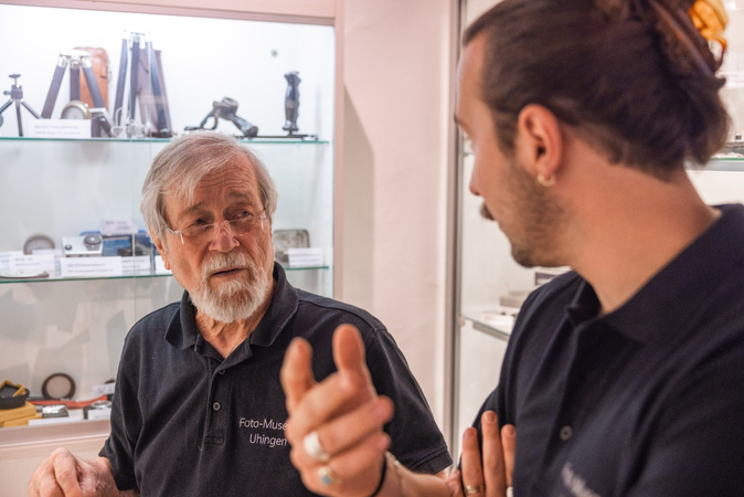 Vor einer ausgeleuchteten Vitrine mit Foto-Zubehör stehen zwei Männer und unterhalten sich gestenreich.