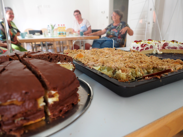 Im Vordergrund sind ein Schoko-, ein Streusel- und ein Erdbeer-Sahne-Kuchen zu sehen. Im Hintergrund sitzen drei Frauen, die unscharf sind, an einem Tisch.