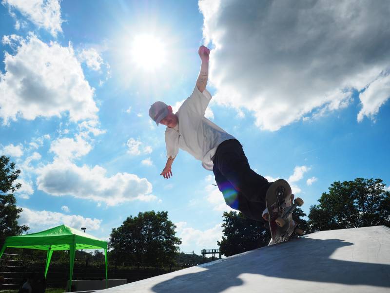 Ein junger Mann mit Schnauzbart steht mit seinem Skateboard an eienr Rampe des Skateparks und die Nase des Skateboards zeigt in den blau-weißen Himmel. Der Junge Mann breitet die Arme aus, um das Gleichgewicht zu halten.
