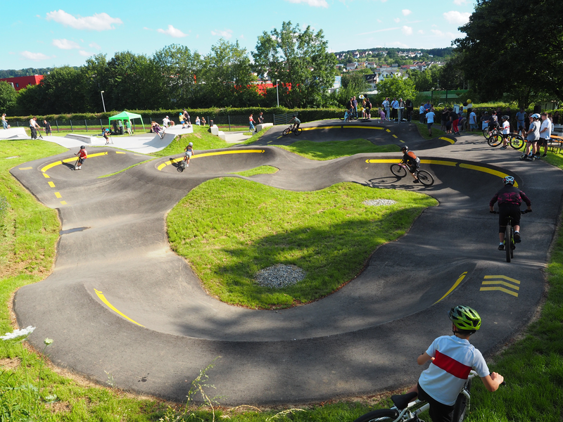 Auf dem asphaltierten und mit gelben Markierungen versehenden Parcours des Pumptracks fahren Kinder und Jugendliche auf ihren Fahrrädern und Rollern. Am Rand halten sich Erwachsene und andere Kinder und Jugendliche auf und schauen zu.