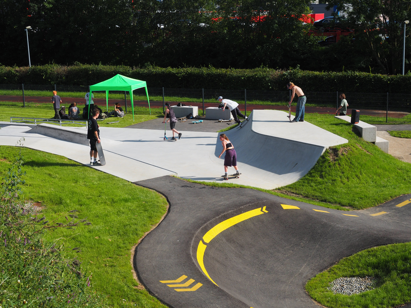 Auf dem Skatepark fahren Jugendliche und junge Erwachsene mit Skateboards oder Rollern. Manche sausen eine Rampe hinab, andere schauen zu.