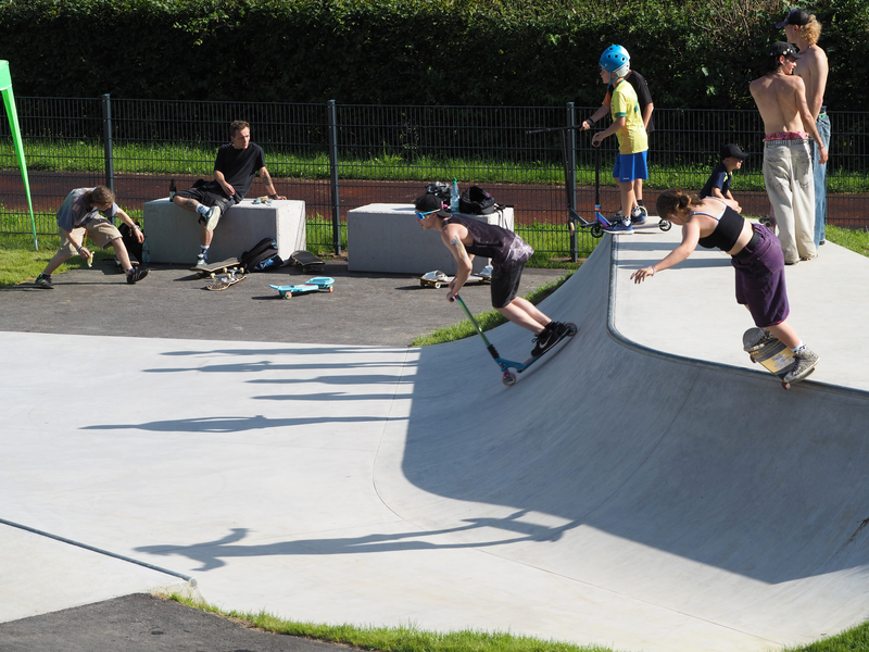 Auf einer Rampe des Skateparks Uhingen stehen mehrere Kinder und Jugendliche. Ein junger Mann rollt mit seinem Roller die Schräge hinab, eine junge Frau rutscht mit ihrem Skateboard die Kante entlang. Im Hintergrund halten sich andere auf Beton-Sitzklätzen auf, Skateboards liegen vor ihnen.