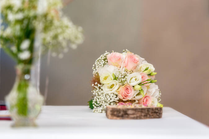 Blumen in einer Glasvase und der Brautstrauß mit rosa-farbenen und weißen Blüten auf einem Tisch mit weißem Tuch.