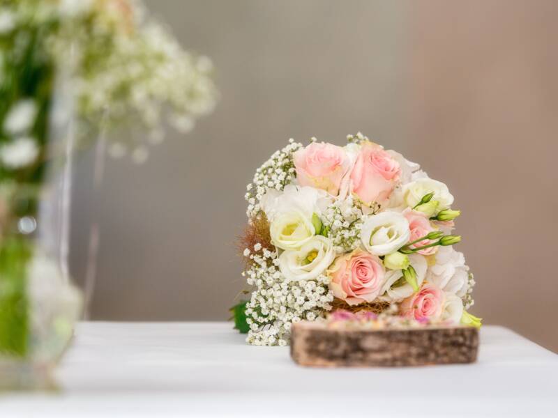 Blumen in einer Glasvase und der Brautstrauß mit rosa-farbenen und weißen Blüten auf einem Tisch mit weißem Tuch.