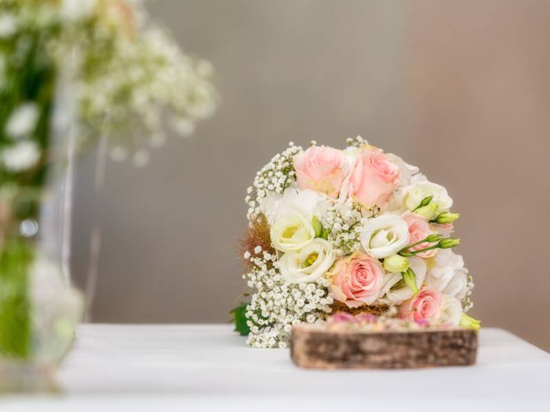 Blumen in einer Glasvase und der Brautstrauß mit rosa-farbenen und weißen Blüten auf einem Tisch mit weißem Tuch.