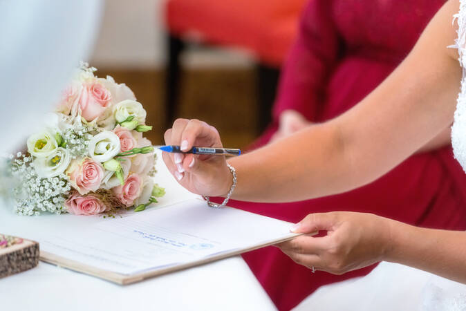 Die Braut ist im Ansatz zu sehen, wie sie einen Stift hält, am Handgelenk des ärmellosen Arms hängt ein Kettchen. Sie unterschreibt auf einem Dokument, das auf dem Tisch liegt - daneben der Brautstrauß.