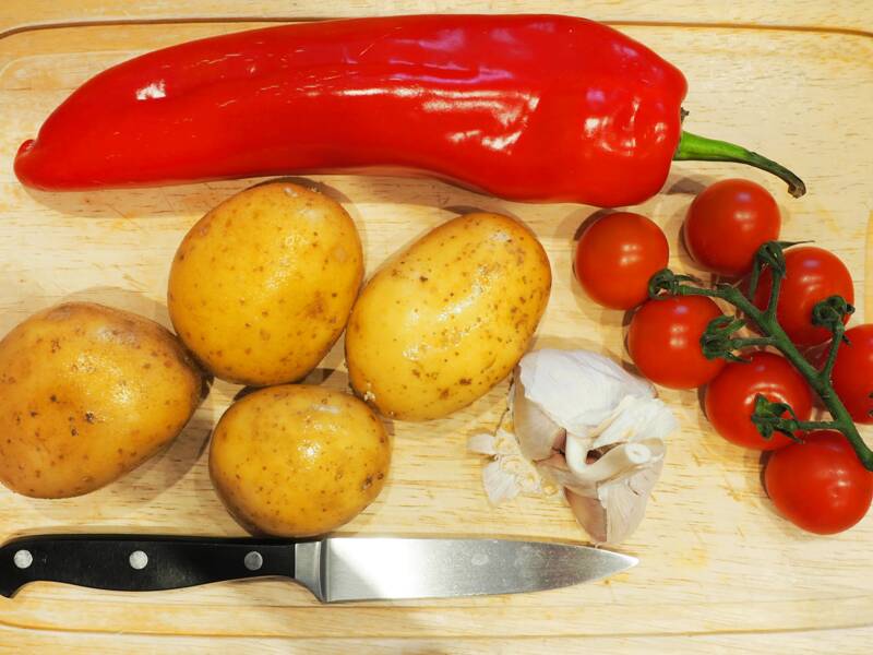 Auf einem Schneidebrett liegen ein Messer, vier Kartoffeln, Knoblauch, eine längliche rote Paprika und Mini-Strauchtomaten.