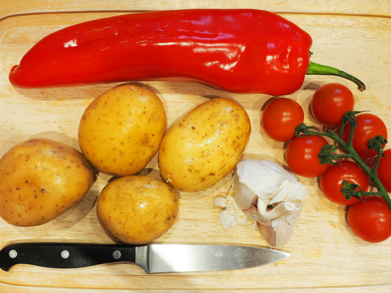 Auf einem Schneidebrett liegen ein Messer, vier Kartoffeln, Knoblauch, eine längliche rote Paprika und Mini-Strauchtomaten.