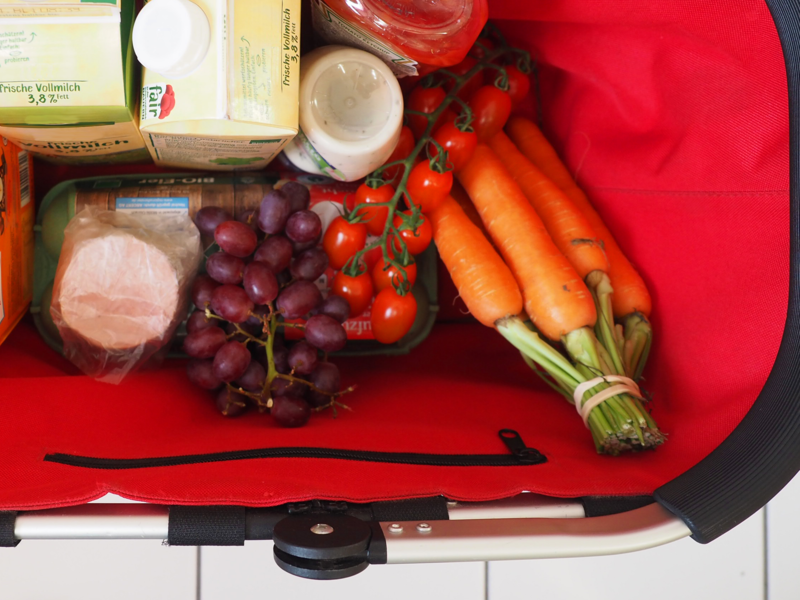 In einem roten Einkaufskorb befinden sich Lebensmittel: 2 Packungen Milch, Süßigkeiten in einer orangefarbenen Packung, rote Weintrauben, Karotten, kleine Strauchtomaten, Ketchup- und Mayonnaise-Tuben, eine Packung Eier und eine Scheibe Leberwurst.