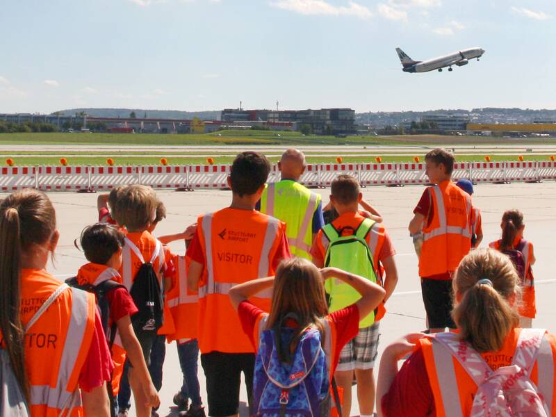 Kinder und Jugendliche stehen, mit orangefarbenen Westen am Rollfeld des Stuttgarter Flughafens und sind von hinten zu sehen. Sie sehen einem Flugzeug zu, das gerade abhebt.