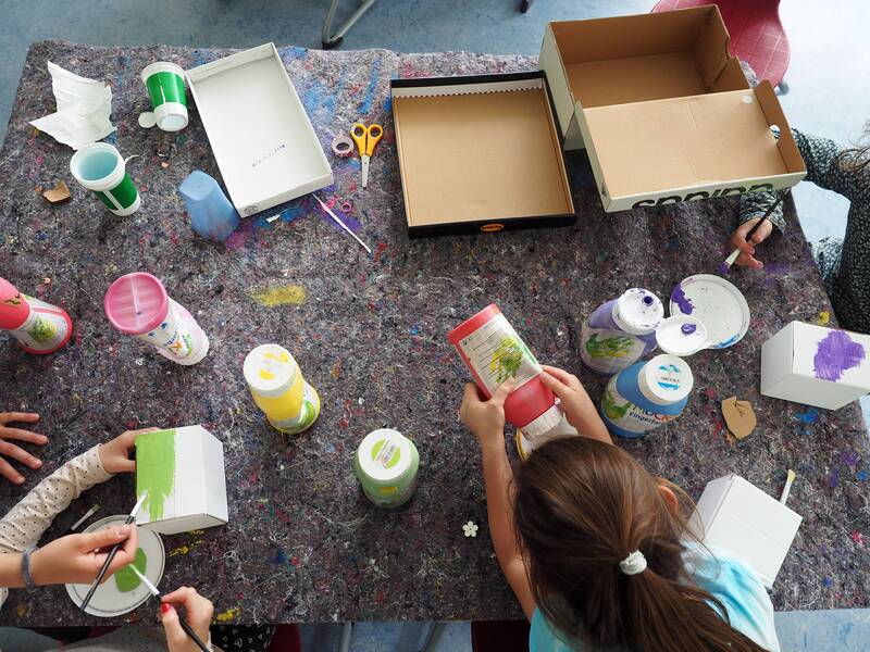 Auf einem Tisch, ausgelegt mit Malerflies als Schutz vor Farbkleksern, sind viele Tuben mit Farben. Kinderhände sind zu sehen, wie sie Pinsel halten und Kartons anmalen oder Farbtuben ausdrücken.