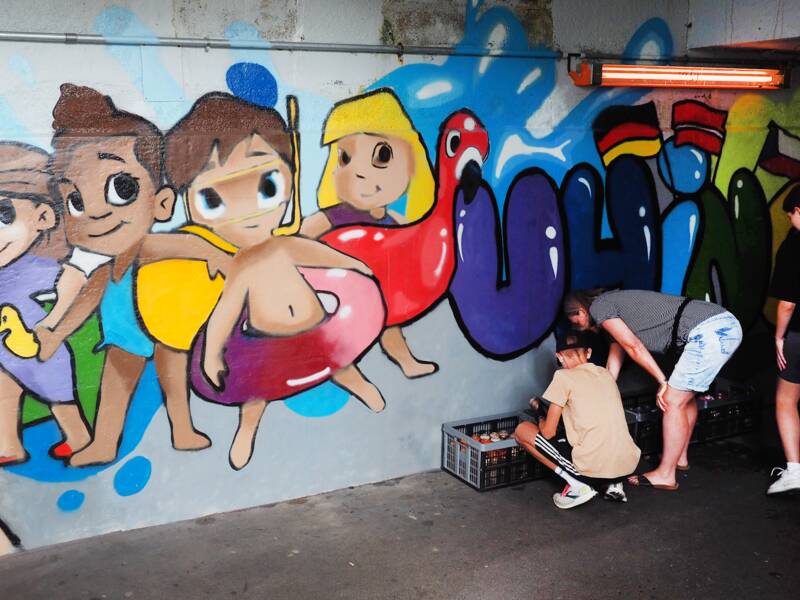 Schüler besprühen für eine Kunstaktion einer Schule die Wand einer Unterführung beim Bahnhof mit Bilder. Auf einem Motiv sind Kinder in Badebekleidung und mit Schwimmringen sowie der angedeutete Schriftzug "Uhingen" mit unterschiedlichen Staatsfahnen.