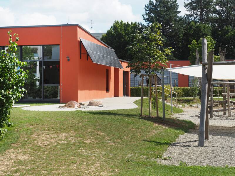 Ein Flachdachbau, der Kindergarten Mittlere Mühle, mit roter Fassade und großen Glasfenstern. Davor eine grüne Wiese und Spielplatz mit Kletterturm und großem Sandkasten.