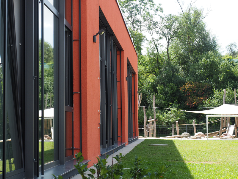 Ein Flachdachbau, der Kindergarten Mittlere Mühle, mit roter Fassade und großen Glasfenstern. Davor eine grüne Wiese mit Spielplatz im Hintergrund.