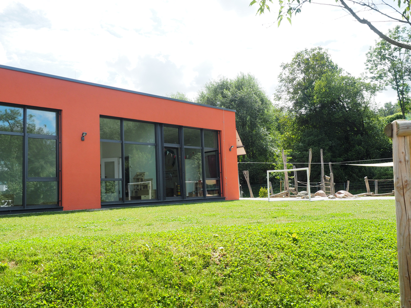 Ein Flachdachbau, der Kindergarten Mittlere Mühle, mit roter Fassade und großen Glasfenstern. Davor eine grüne Wiese mit Spielplatz im Hintergrund.