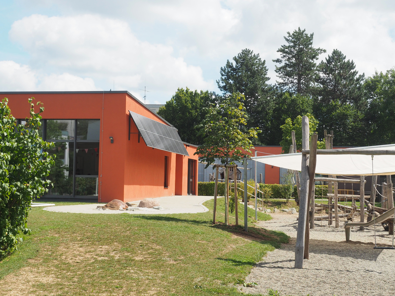 Ein Flachdachbau, der Kindergarten Mittlere Mühle, mit roter Fassade und großen Glasfenstern. Davor eine grüne Wiese und Spielplatz mit Kletterturm und großem Sandkasten.