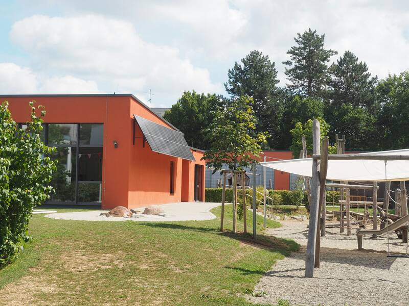 Ein Flachdachbau, der Kindergarten Mittlere Mühle, mit roter Fassade und großen Glasfenstern. Davor eine grüne Wiese und Spielplatz mit Kletterturm und großem Sandkasten.