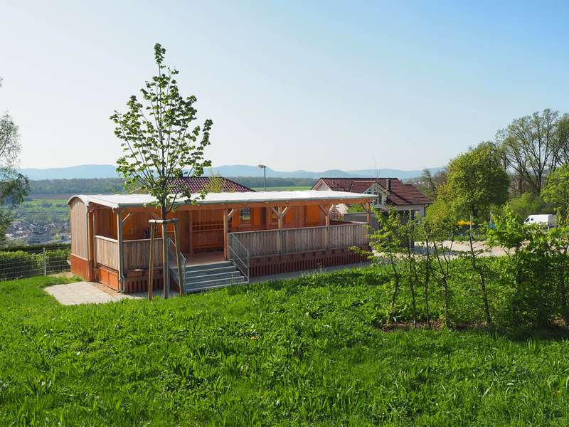 Vor einer Rasenfläche ist der Naturkindergarten: ein flacher Holzbau, der aus einem umgebauten Bauwagen sowie einer überdachten Holzterrasse besteht. Auf der Wiese stehen einige junge, bis zu drei Meter hohe Bäume, im Hintergrund erstreckt sich das Mittelgebirge der Schwäbischen Alb vor dem blauen Himmel.