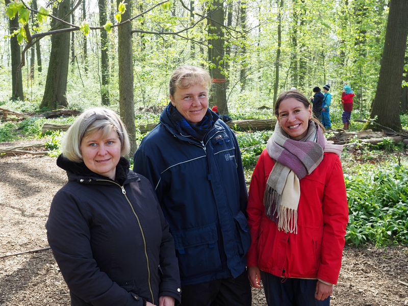 Drei Frauen, das Team des Naturkindergartens, steht im Wald, umgeben von Bäumen, Sträuchern und anderen Pflanzen. Im Hintergrund spielen Kinder mit Stöcken.