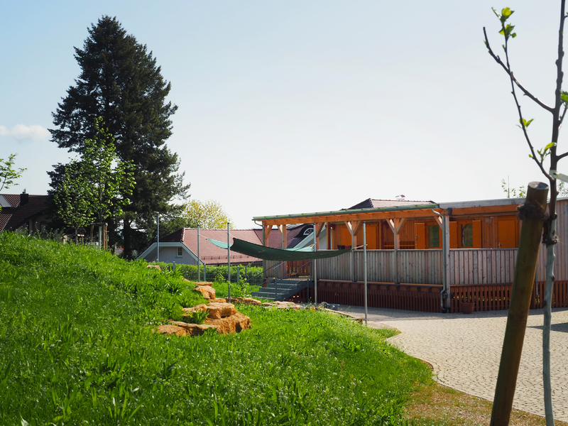 Vor einer Rasenfläche ist der Naturkindergarten: ein flacher Holzbau, der aus einem umgebauten Bauwagen sowie einer überdachten Holzterrasse besteht. Vor dem Gebäude ist an Eisenpfählen ein Baldachin als Sonnenschutz aufgespannt. Auf der Wiese stehen einige junge, bis zu drei Meter hohe Bäume, im Hintergrund erstreckt sich das Mittelgebirge der Schwäbischen Alb vor dem blauen Himmel.