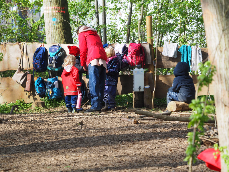 An einem Holzzaun, bestehend aus Pfosten und zwei dicken Brettern, hängen im Wald des Naturkindergartens mehrere Rucksäcke neben- und übereinander. Davor stehen Kinder und eine Erzieherin auf dem mit Holzspänen ausgelegten Waldboden.