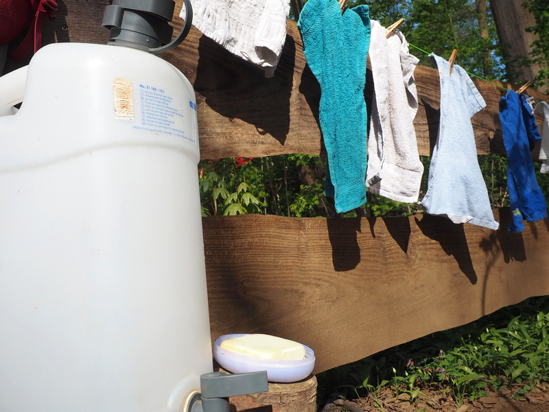 Auf einem Baumstamm in einem Waldstück steht ein Wasserkanister mit einer Öffnung unten, daneben ist ein Seifenschälchen mit Seife. So können die Kinder Hände waschen. An einem Holzzaun daneben hängen an einer Leine kleine Handtücher zum Abtrocknen.