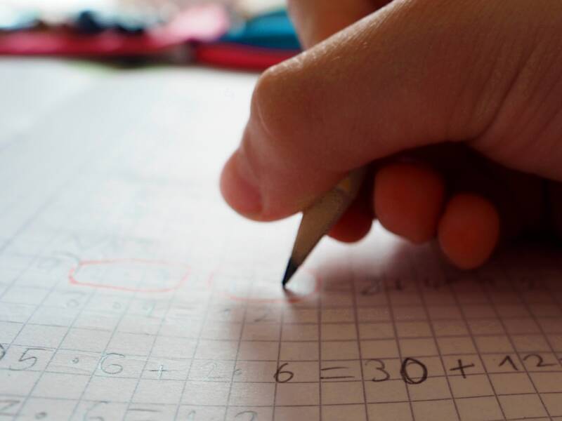 Eine Kinderhand hält einen Buntstift mit blauer Mine und schreibt in ein kleinkariertes Matheheft Zahlen zu einer Rechenaufgabe.
